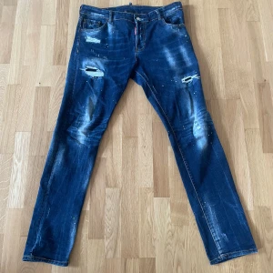 Dsquared2 blå jeans - Säljer ett par blå jeans från Dsquared2 med coola slitningar och färgstänk på både fram- och baksida. Jeansen har klassisk femficksdesign, rak passform och en snygg Dsquared2-patch bak i midjan. Perfekt för dig som gillar en edgy streetstil.