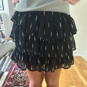 Svart volangkjol med blixtmönster - Supersnygg svart kortkjol med flera lager volanger och coolt vitt blixtmönster. Kjolen är luftig och har en skön passform som sitter bra i midjan. Storleken är 176 men sitter perfekt på mig som är S! Prislappen är kvar, aldrig använd och nypris är 600 kr. Perfekt till alla plagg och årstider!🌷💓 Jag kan även förhandla om priset