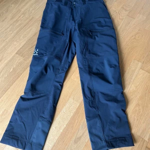 Svarta täckbyxor från Haglöfs dam - Haglöfs Rugged Relaxed Pant Women, storlek 40(S)/US 10(S), i färgen True Black. Byxorna har flera fickor med dragkedja och justerbara benslut med tryckknappar. Modell short vilket innebär något kortare benlängd. Använda en gång som nya. 