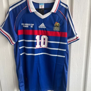 Frankrike Zidane VM 98 fotbollströja - Säljer en ikonisk blå Frankrike fotbollströja från VM 98 med nummer 10 och Zidane på ryggen. Adidas-logga, broderat landslagsmärke och vita/röda detaljer. Ränder över bröstet och klassisk krage. Perfekt för dig som älskar retro och fotbollshistoria.