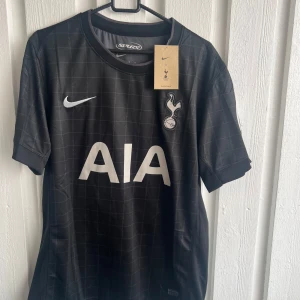 Tottenham svart matchtröja Bergvall #15 - Snygg svart Tottenham Hotspur fotbollströja från Nike med diskret rutmönster, vita tryck och Bergvall #15 på ryggen. Kortärmad modell med klubbmärke, sponsorlogga och Premier League-detaljer på ärmen. Perfekt för dig som vill representera Spurs.