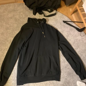 Svart hoodie med huva och ficka - Svart hoodie med klassisk huva och snörning, samt en stor magficka framtill. Tröjan har långa ärmar och ribbade muddar vid ärmslut och nederkant. Perfekt för chill dagar eller när du vill ha en enkel och stilren look.