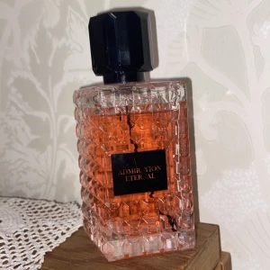 Admiration Eternal Eau de Parfum 100ml - Säljer en parfymflaska Admiration Eternal från Coscentra, Eau de Parfum 100ml. Flaskan är fyrkantig med ett coolt rutmönster i transparent glas och har en svart, kantig kork. Innehållet är orangefärgat och flaskan känns lyxig och modern.