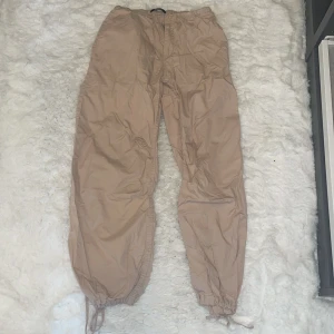 Beige cargopants med snörning - Säljer ett par beige cargopants med elastisk midja och snörning vid bensluten. Byxorna har en loose passform och är tillverkade i ett lätt syntetmaterial, perfekt för chill dagar. Färgen är neutral och passar till det mesta. Sköna att bära och enkla att styla. Från Ginatricot  