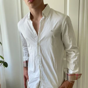 Burberry skjorta vit - Burberry skjorta vit i väldigt bra skick förutom en fläck på ryggen som man ser bäst på bild 5. Har ej provat om den går bort men tror den borde göra det. Kan gå ned i pris vid snabb affär. Modellen bär M och är 180,75 kg. Kom md frågor! 