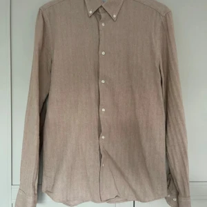 Beige skjorta från Bläck i bomull - Snygg beige skjorta från Bläck i storlek S med skräddarsydd passform. Skjortan har ett diskret fiskbensmönster, klassisk krage med knappar och vita knappdetaljer framtill. Perfekt för dig som gillar stilrena och neutrala plagg.