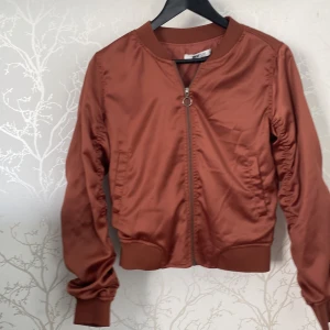 Rostbrun bomberjacka Gina Tricot - Snygg rostbrun bomberjacka från Gina Tricot i storlek 36. Jackan har en rund hals, dragkedja med ringdetalj och rynkade sömmar längs ärmar och rygg. Tillverkad i blankt syntetmaterial med ribbade muddar vid ärmslut och nederkant. Perfekt för dig som gillar streetstyle.