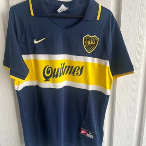 Boca Juniors Maradona tröja Nike M - Säljer en Boca Juniors fotbollströja med Maradona #10 på ryggen. Tröjan är marinblå med breda gula och vita ränder över bröstet, klassisk krage och tryckta loggor. Materialet är lätt polyester, perfekt för match eller träning. Snygg CABJ-emblem och Quilmes sponsortryck.