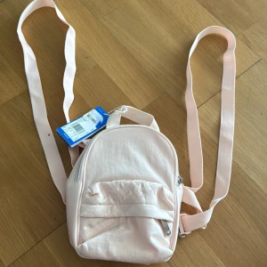 Mini rosa ryggsäck från Adidas - oanvänd  - Säljer en söt mini ryggsäck från Adidas i ljusrosa färg. Väskan har justerbara axelremmar, ett litet ytterfack med dragkedja och diskret Adidas-logga framtill. Materialet är mjukt och lätt polyester, perfekt för att bära småsaker. Supersnygg att ha till vardags!