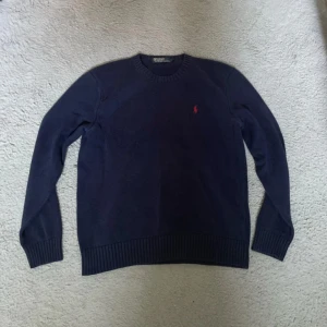 Mörkblå stickad tröja Polo Ralph Lauren - Snygg mörkblå stickad tröja från Polo Ralph Lauren i storlek S. Klassisk rund hals och ribbade muddar. På bröstet finns den ikoniska röda Polo-loggan. Perfekt för dig som gillar stilrena och tidlösa plagg med premiumkänsla.