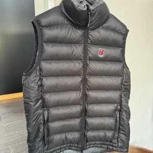 Svart dunväst från Fjällräven - Sjukt snygg svart dunväst från Fjällräven med medelhög krage och quiltat dun. Västen har en dubbeldragkedja framtill och Fjällräven-logga på bröstet. Perfekt för lager-på-lager och chill höstdagar. Materialet är lätt och smidigt, samtidigt varmande och stilrent. Den är använd 1 gång tidigare. Priset kan diskuteras, ordinarie pris låg på 2000kr