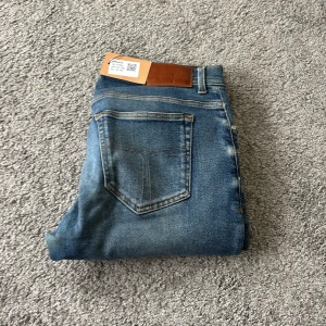 Blå jeans Tiger of Sweden 33/34 - Snygga blå jeans från Tiger of Sweden i storlek 33/34. Modellen har klassisk femficksdesign, raka ben och en brun läderpatch med logga bak i midjan. Jeansen är tillverkade i twillvävt denim och har en fräsch tvätt med lätt slitning på låren.
