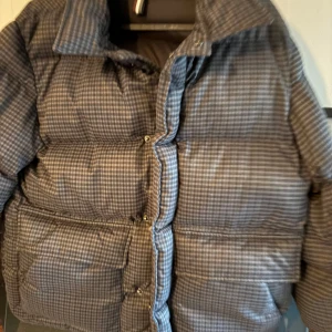Grå rutig pufferjacka från Colmar - Snygg pufferjacka från Colmar i grått med rutigt mönster. Jackan har hög krage, stora fickor och knappar framtill. Perfekt för dig som vill ha en varm och trendig jacka med streetkänsla. Colmar-logga på ärmen ger extra stilpoäng. Ny pris ca 5 6 tusen 