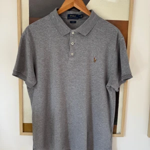 Grå slim fit polo från Ralph Lauren, storlek L - Snygg grå pikétröja från Ralph Lauren i slim fit-modell. Klassisk krage, tre vita knappar och broderad logga på bröstet. Tillverkad i mjuk bomull som känns skön mot huden. Perfekt för dig som gillar stilrena och tidlösa plagg.
