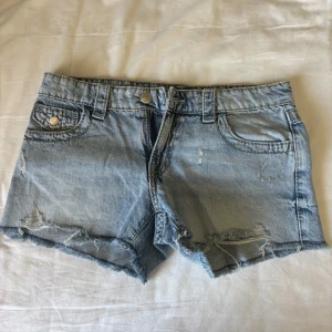 Jeansshorts med snygga fickor  - Snygga ljusblå jeansshorts med fransig kant och slitningar från Gina Tricot. Baksidan har två fickor med detaljerad broderad paisleymönster och metallknappar. Klassisk femficksmodell och normal passform, perfekta för varma dagar. De har ingen prislapp men är aldrig använda. Stl 170 passar mig som har xs/s i byxor.