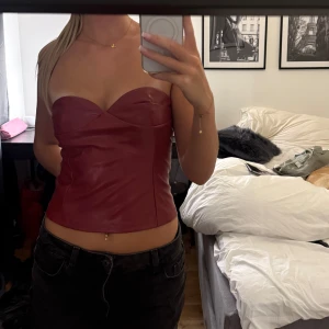 Mörkröd skinnkorsett med sweetheart - Säljer en mörkröd korsett-topp i skinnimitation med snygg sweetheart-urringning. Toppen är axelbandslös och har en tight passform som framhäver midjan. Perfekt för dig som vill ha en edgy och trendig look. Passar till både jeans och kjol för en cool vibe.