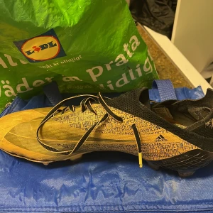 Adidas gula och svarta fotbollsskor - Säljer ett par Adidas fotbollsskor i storlek 43 1/3 med coolt textmönster i gult och svart. Skorna har snörning, platt sula med dobbar och en snygg detalj på hälen. Perfekt för dig som vill sticka ut på planen med stil.