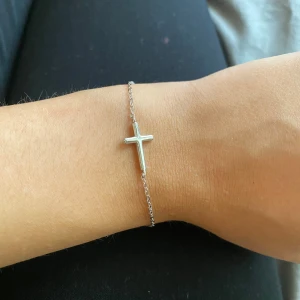 Silverarmband med kors från e&e London - Stilrent armband i äkta silver från e&e London med ett minimalistiskt kors som hänger på en tunn kedja.      Aldrig andvändt och perfekt för dig som gillar enkel och snygg design. Lätt att matcha med andra smycken och har en smidig karbinlås. Levereras med originalpåse.      Köpt i London för drygt 600kr.
