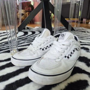 Vans Rowley Classic 66/99 (42) - Säljer ett par Vans Rowley sneakers i vitt med klassiskt rutigt mönster på sidan. Skorna har snörning, platt sula och rund tå. Ovandelen är i slitstark skinn och textil, med svart och vit färgdetalj. Perfekta för skate och streetstyle. Sulan är gummibrun med ikoniskt mönster.