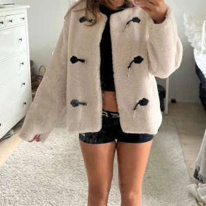 Beige teddyjacka med svarta detaljer - Säljer en helt oanvänd beige skitsnygg teddyjacka från Gina Tricot med coola svarta snörningsdetaljer framtill. Jackan har lång ärm, rund hals och är gjord i mjukt teddy material. Perfekt till höst/vinter åren! 