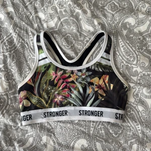 Blommig sporttopp från Stronger XS - Sporttopp från Stronger i storlek XS med tropiskt blommigt mönster. Elastisk vit kant med logga runt om. Materialet är 73% polyester och 27% spandex, vilket gör den stretchig och bekväm för träning. Lät AI beskriva, men har bara använt 3 gånger typ och den är jätte gullig🥰(pris kan diskuteras)