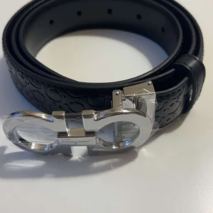 Svart bälte/plånbok från Salvatore Ferragamo - Snyggt svart bälte från Salvatore Ferragamo med präglat mönster längs hela remmen och en stilren, silverfärgad spänne i metall. Bältet är tillverkat i exklusivt skinn och har en modern, elegant design som lyfter din outfit.