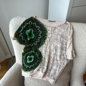 Desigual t-shirt med färgglatt mönster - Unik t-shirt från Desigual med korta ärmar och rund hals. Ena sidan har ett stort, färgstarkt blommigt mönster i grönt, blått och rött, medan andra sidan är ljusbeige med svarta snirkliga linjer. Materialet känns mjukt och lätt, perfekt för dig som gillar statement-plagg. Baksidan är beige utan detaljer. 