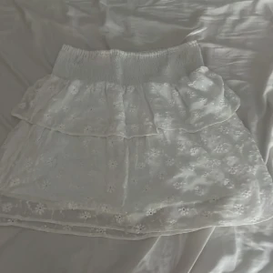 Vit kortkjol med broderade blommor - Supersöt vit kortkjol med bred resår i midjan och dubbla lager. Kjolen har broderade blommor över hela tyget och ett tunt, luftigt material med innerfoder. Perfekt för dig som gillar romantisk och feminin stil. Passar till sneakers eller sandaler.