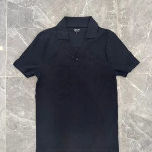 Svart ribbad t-shirt från ASOS - Svart ribbad t-shirt från ASOS med klassisk krage och v-ringning med knapp. T-shirten har korta ärmar och är gjord i mjuk bomull som känns skön mot huden. Perfekt för dig som gillar stilrena och enkla plagg med en twist.