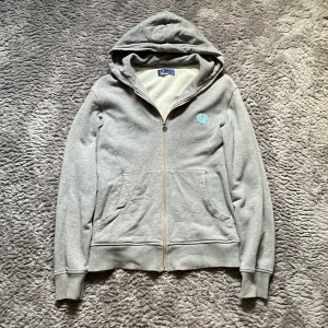Grå hoodie från Fred Perry med dragkedja - Säljer en grå hoodie från Fred Perry i storlek S. Tröjan har huva, två fickor fram och en guldfärgad dragkedja. På bröstet sitter Fred Perrys klassiska blå logotyp. Materialet är mjukt och känns som bomull, perfekt för chill dagar.