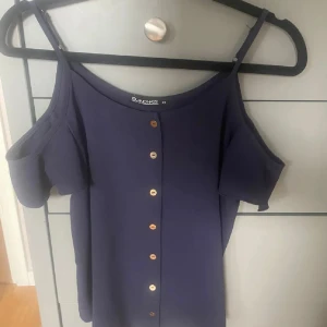 Mörkblå offshoulder topp - Snygg mörkblå topp från Island Haze med offshoulder-design och smala axelband. Framtill sitter dekorativa knappar i guldton som ger en cool touch. Perfekt för dig som gillar stilrena och trendiga plagg med lite extra edge.