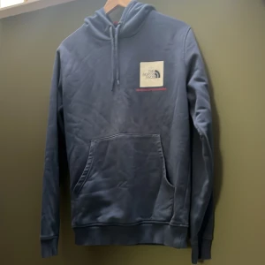 Blå hoodie från The North Face - Säljer en snygg blå hoodie från The North Face med klassisk logga på bröstet och ryggen. Tröjan har huva med snörning, stor magficka och långa ärmar. Perfekt för chill dagar. Materialet är mjuk bomull och polyesterblandning.
