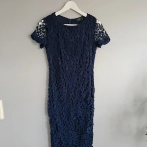 Marinblå spetsklänning AX Paris - Säljer en marinblå spetsklänning från AX Paris med korta ärmar och rund hals. Klänningen har ett vackert blommigt spetsmönster och är fodrad med mjukt tyg. Passformen är normal och längden går till knäna. Perfekt för dig som gillar eleganta detaljer och stilren look.