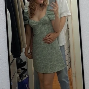 Grön blommig kortklänning med puffärm - Supersöt ljusgrön kortklänning med små vita blommor, puffärmar och fyrkantig urringning. Klänningen har en figurnära passform upptill och lätt utsvängd kjol. Perfekt för varma dagar och har en mjuk, skön känsla. Materialet känns som bomull eller polyester.