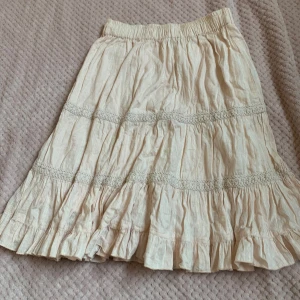 Beige volangkjol med spetsdetaljer - Säljer en beige kjol med volanger och fina spetsdetaljer. Kjolen har resår i midjan och är tillverkad i ett lätt material som ger ett luftigt intryck. Perfekt för dig som gillar en romantisk och bohemisk stil.