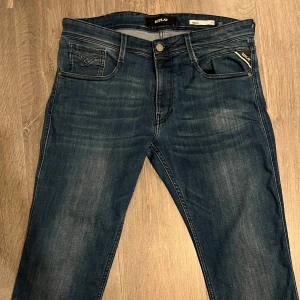 Replay Anbass jeans - Säljer ett par mörkblå Replay Anbass jeans med klassisk femficksdesign och snygga slitningar. Jeansen har normal passform och raka ben, med diskreta detaljer och logga på myntfickan. Perfekta för dig som gillar stilrena jeans med lite tvättad look.