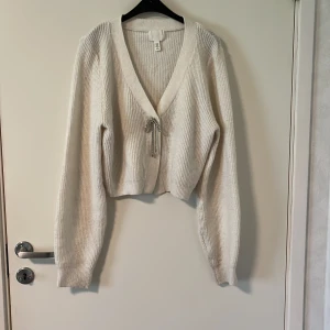 Vit croppad kofta från H&M - Säljer en vit, stickad kofta från H&M med croppad passform. Koftan har långa ärmar, ribbad struktur och fin roset framtill. Perfekt för lager-på-lager och enkel att styla till olika outfits.
