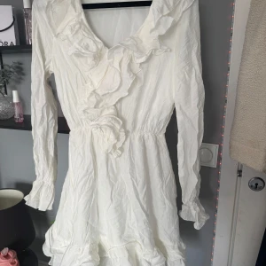 Vit volangklänning från SHEIN - Säljer en söt vit klänning från SHEIN i storlek XS. Klänningen har långa ärmar, volangdetaljer längs ringningen och framtill samt en lätt rynkad midja. 