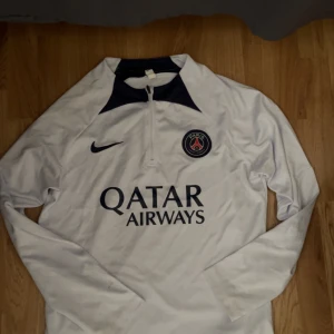 Vit långärmad PSG fotbollströja från Nike - Säljer en vit långärmad Paris Saint-Germain fotbollströja från Nike med klubbmärke på bröstet och Qatar Airways som sponsortryck. Tröjan har dragkedja vid halsen och mörkblå detaljer vid kragen. Perfekt för dig som älskar PSG!