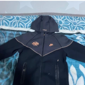 Svart hoodiejacka från Nike - Säljer en svart hoodiejacka från Nike med dragkedja och huva. Jackan har orange Nike-logga och grafiskt tryck på bröstet samt två fickor med dragkedja. Snygga kontrastsömmar och sportig design.