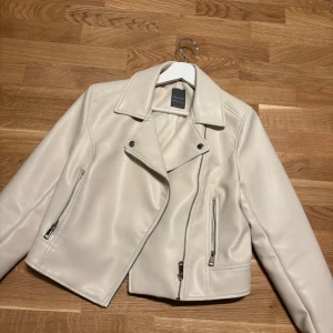 Vit skinnjacka från Primark - Snygg vit skinnjacka med klassisk bikerstil från Primark. Jackan har silvriga dragkedjor, bred krage och snedstängning framtill. Perfekt för att lyfta vilken outfit som helst.