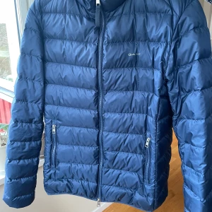 Blå pufferjacka från Gant - Säljer en blå lätt pufferjacka från Gant med dragkedja framtill och två fickor med dragkedja. Jackan har lång ärm och quiltad design, perfekt för kyligare vår/sommar/höst dagar. Enkel och stilren modell med diskret logga på bröstet.