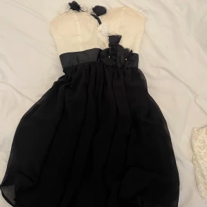 Svart och vit klänning med blommiga detaljer - Säljer en söt klänning med vit överdel och svart kjol. Axelbandet är dekorerat med svarta och vita blommor. Kjolen är luftig och faller fint. Perfekt för dig som vill sticka ut lite extra! Den är aldrig använd men har blivit missfärgad, vet dock inte av vad eller om det går bort därför säljer jag den billigt 