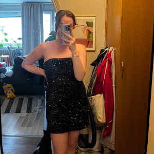 Svart paljettklänning från Lindex - Säljer en svart axelbandslös kortklänning från Lindex, täckt med glittriga paljetter. Klänningen har en hjärtformad urringning och smala axelband, perfekt för att sticka ut på festen.