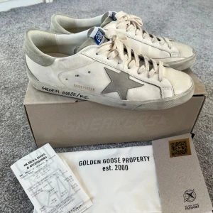 Golden Goose Superstar sneakers med grå stjärna - Golden Goose Superstar sneakers i vitt skinn med grå mockadetaljer och en grå stjärna på sidan. Pga för stor storlek är skorna använda ett fåtal gånger och man kan i princip inte se att de är använda. Med andra ord absolut toppskick! Nypris 5599kr. Kartong, dustbag och kvitto från NK följer med. 