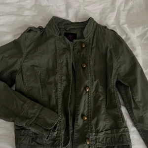 Olivgrön jacka från Ann Christine - Säljer en olivgrön jacka från Ann Christine med snygga knappar framtill och axeldetaljer. Jackan har lång ärm och en rak passform. Perfekt för dig som vill ha en stilren och enkel look.💕