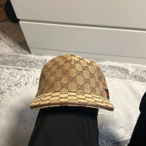 Gucci keps beige - Säljer en beige keps från Gucci med klassiskt GG-monogram över hela kepsen. Kepsen har en böjd skärm och detaljer med grönt och rött band på sidan. Justerbar rem baktill. Perfekt för dig som gillar exklusiva accessoarer. Storlek M.