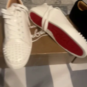 Vita sneakers med nitar och röd sula - Säljer ett par vita sneakers med klassisk röd sula och coola nitar framtill. Skorna har snörning och en stilren design som sticker ut.