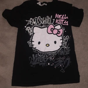Svart Hello Kitty t-shirt från FB Sister - Aldrig använt, svart t-shirt med stort Hello Kitty-tryck framtill och graffitiinspirerad text i vitt och rosa. Klassisk passform med rund halsringning och korta ärmar. Perfekt för dig som gillar söta och lekfulla motiv.