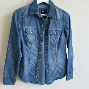 Blå jeansskjorta med prickigt mönster från H&M - Snygg blå jeansskjorta från H&M med små vita prickar och tryckknappar. Skjortan har två bröstfickor med lock och lång ärm med knappar vid manschetten. Perfekt att styla till jeans eller kjol för en cool look.